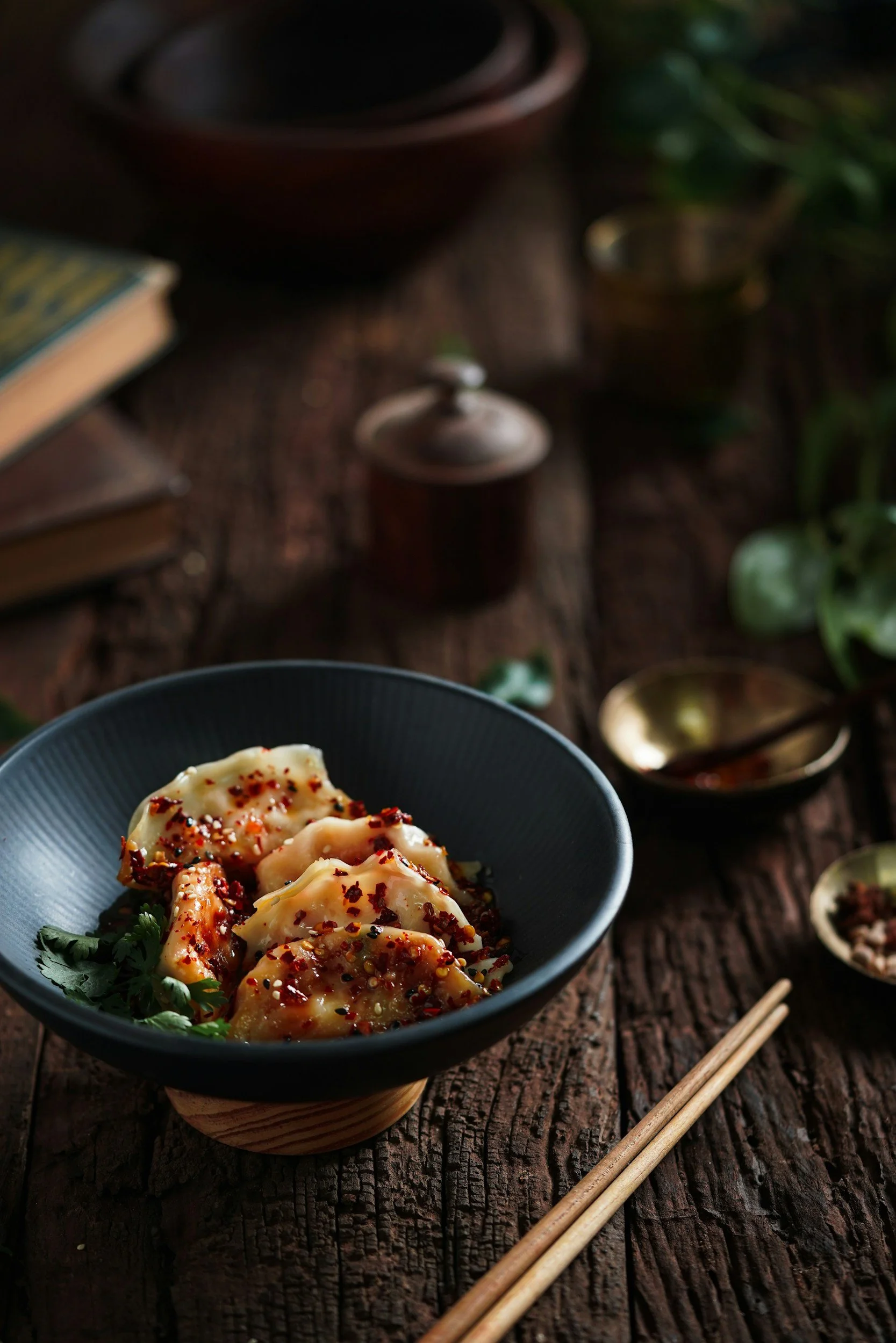 Close-up of a black bowl with dumplings topped with chili flakes and herbs, placed on a rustic wooden surface, with chopsticks nearby and various bowls and plants in the background.