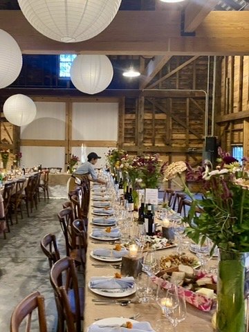 Long dining table set with plates, glasses, and wine bottles, decorated with bouquets of flowers in a rustic wooden venue with hanging paper lanterns.