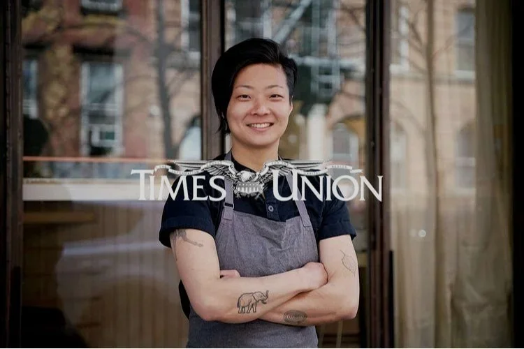 A woman with short black hair, smiling, wearing a dark shirt and apron, standing in front of a glass door with the 'Times Union' logo, and crossing her arms.