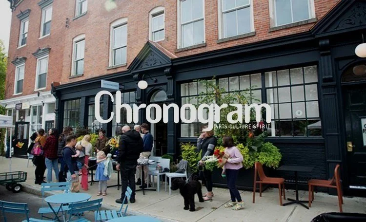 People gather outside a storefront with the sign 'Chronogram Arts Culture Spirit,' some holding flowers, with blue chairs and tables on the sidewalk in an urban setting.