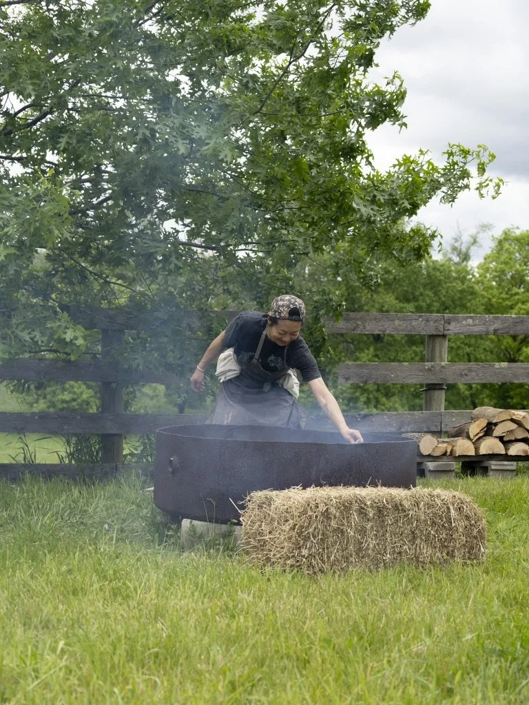Person tending a fire in a large metal fire pit outdoors, surrounded by green grass and trees, with a hay bale in the foreground and a wooden fence in the background.