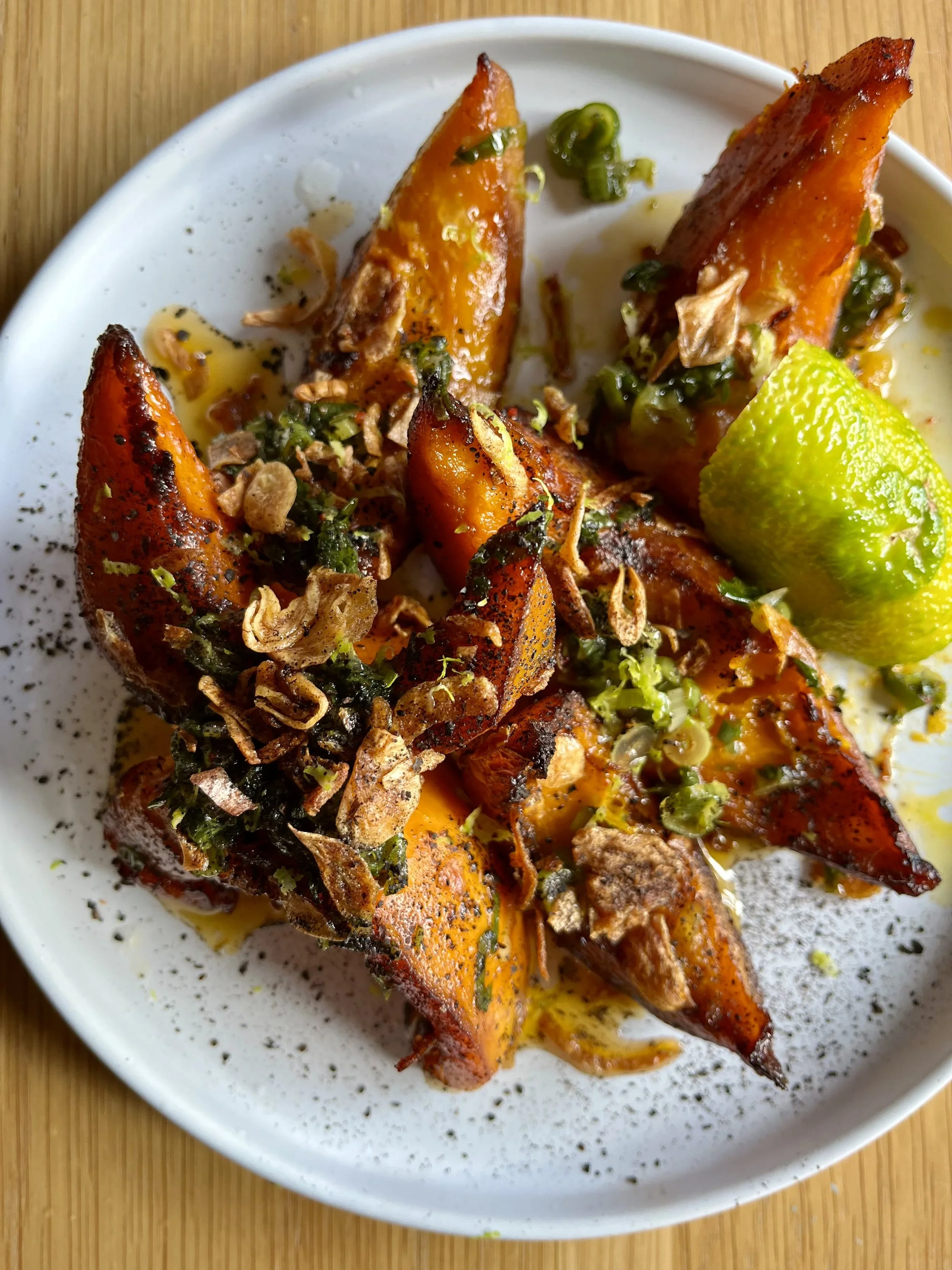 Grilled sweet potato wedges garnished with chopped green onions and fried garlic, served with a lime wedge on a white plate.