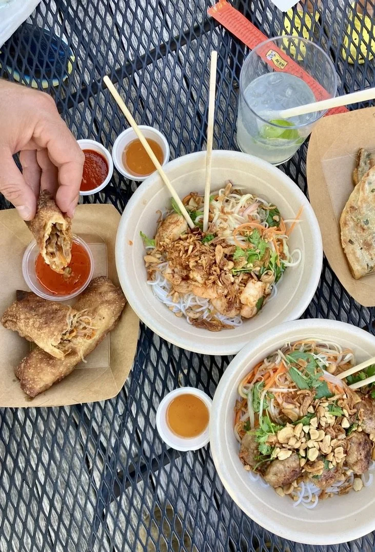Two plates of Vietnamese noodle dishes with herbs, vegetables, and fried shallots, accompanied by spring rolls with dipping sauce, and drinks including a lime-infused soda with a straw and a glass of water with ice and lime.