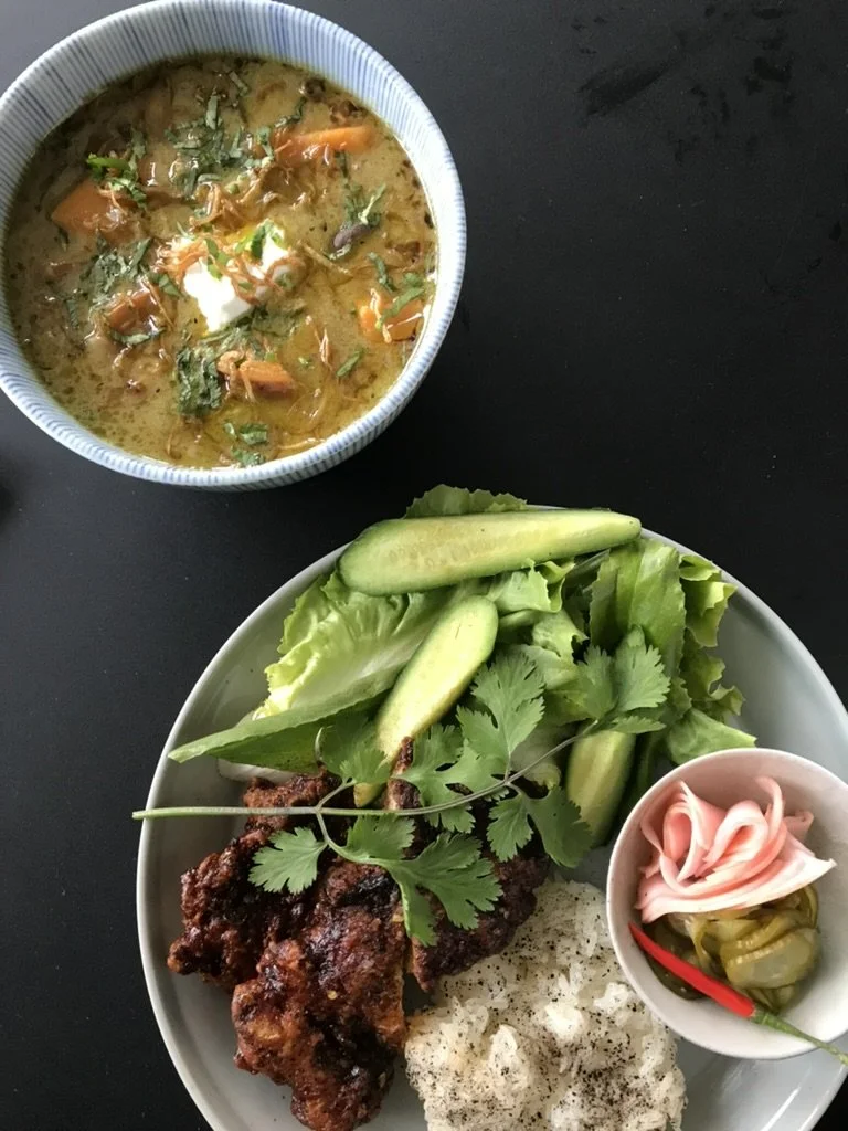 Plate with fried chicken, cucumber slices, fresh cilantro, and green leafy vegetables; small bowl with pickled vegetables and pink ham; bowl of curry with carrots, herbs, and a dollop of white sauce.