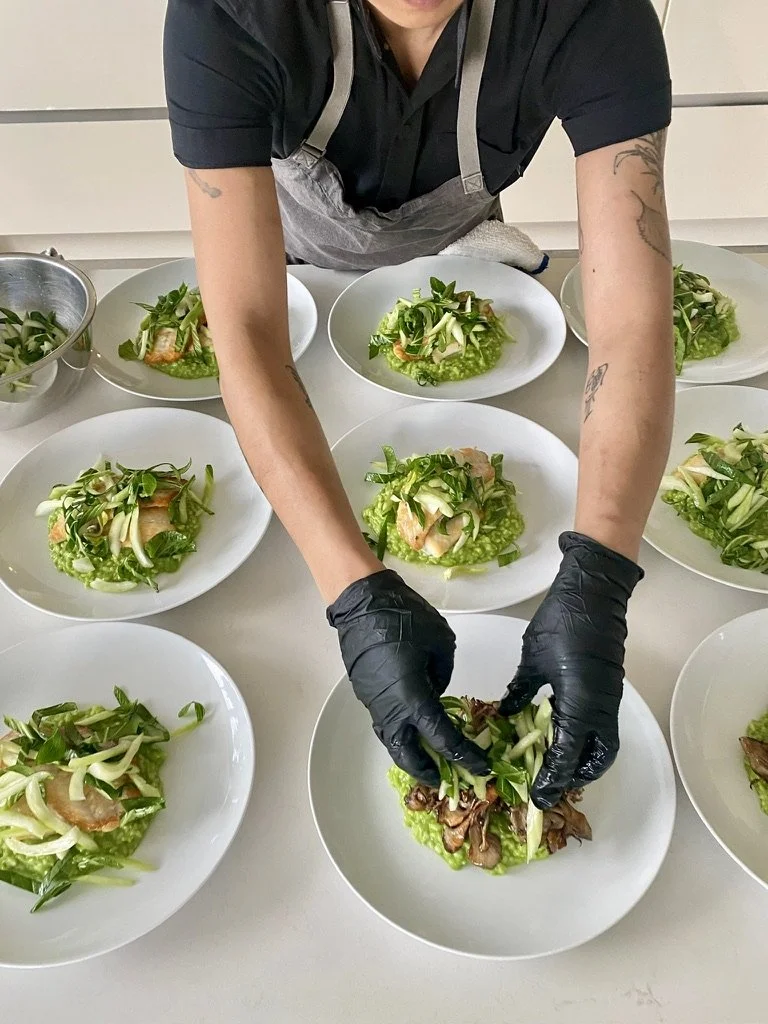 A chef or cook preparing salads on white plates, wearing black gloves and a black shirt, in a professional kitchen or food prep area.