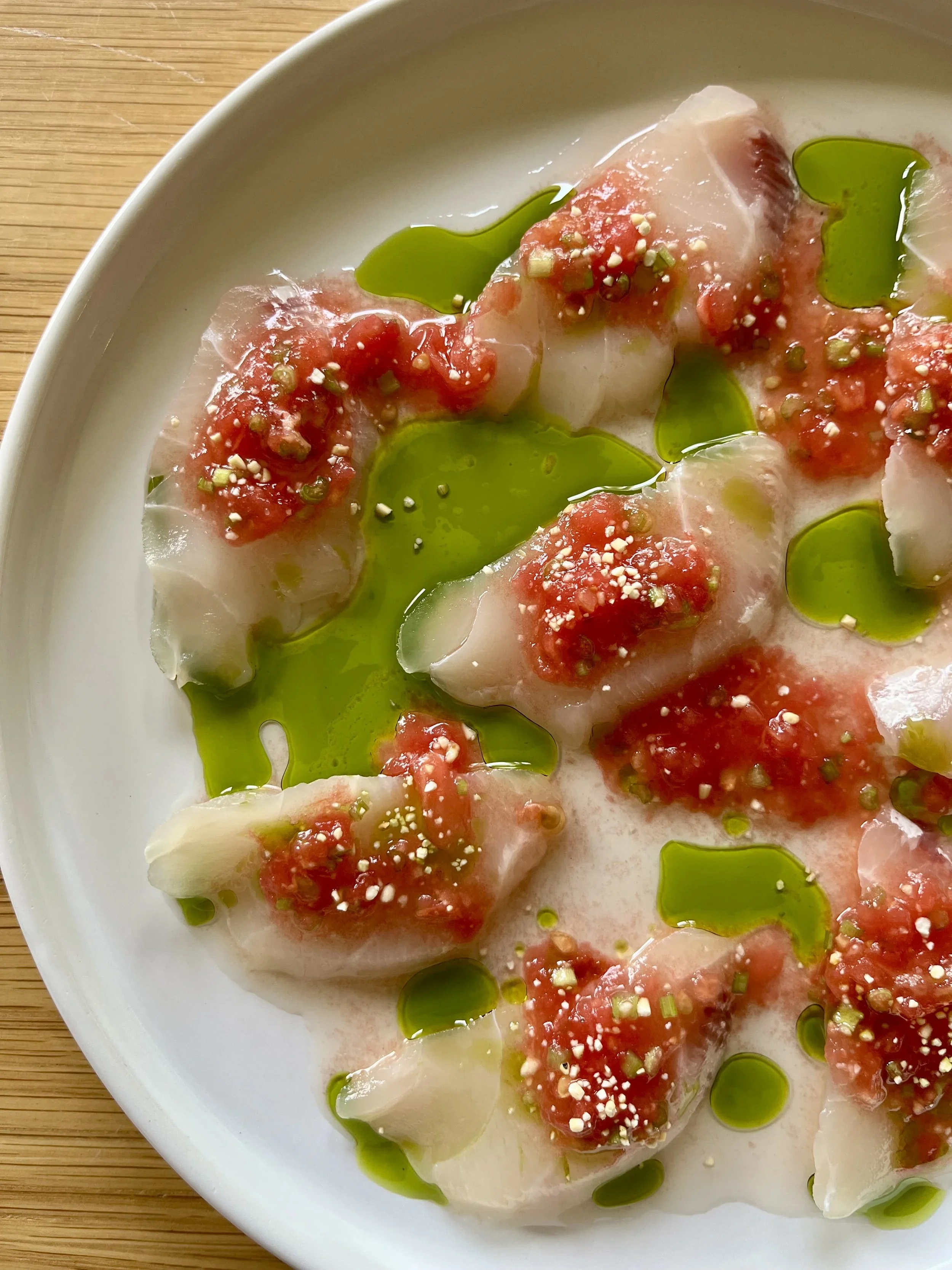 Close-up of raw fish fillets topped with red sauce, sesame seeds, and chopped green onions, drizzled with green oil, resting on a white plate.