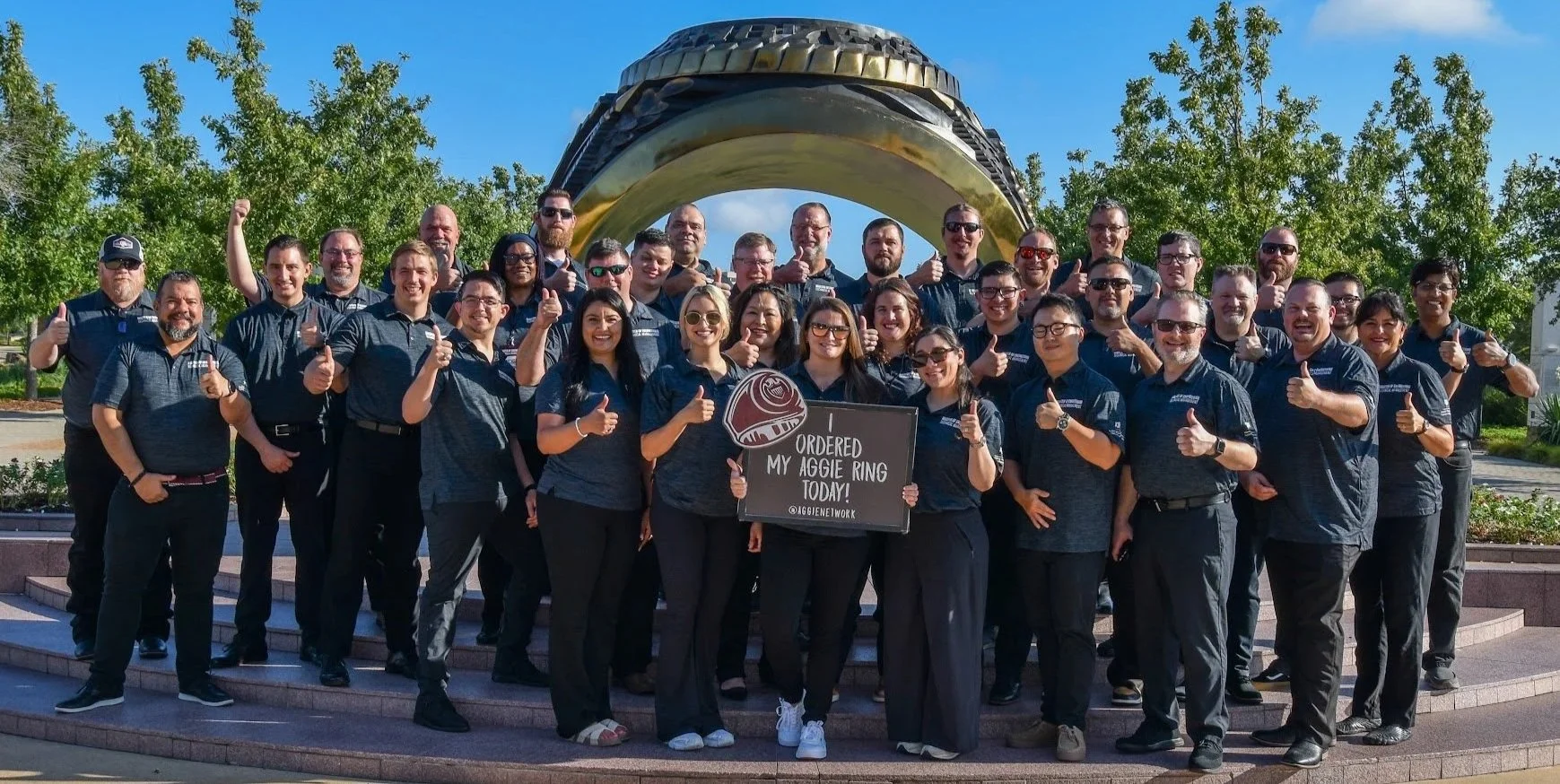Group of people outdoors, wearing matching shirts, standing on steps in front of a large gold-colored sculpture, holding a sign that says "Ordered my Aggie ring today!" and giving thumbs up.
