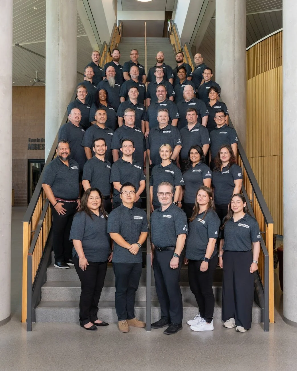 Group photo of 31 people, mostly in matching dark blue polo shirts, standing on a staircase in a modern building with wooden and concrete architectural features.