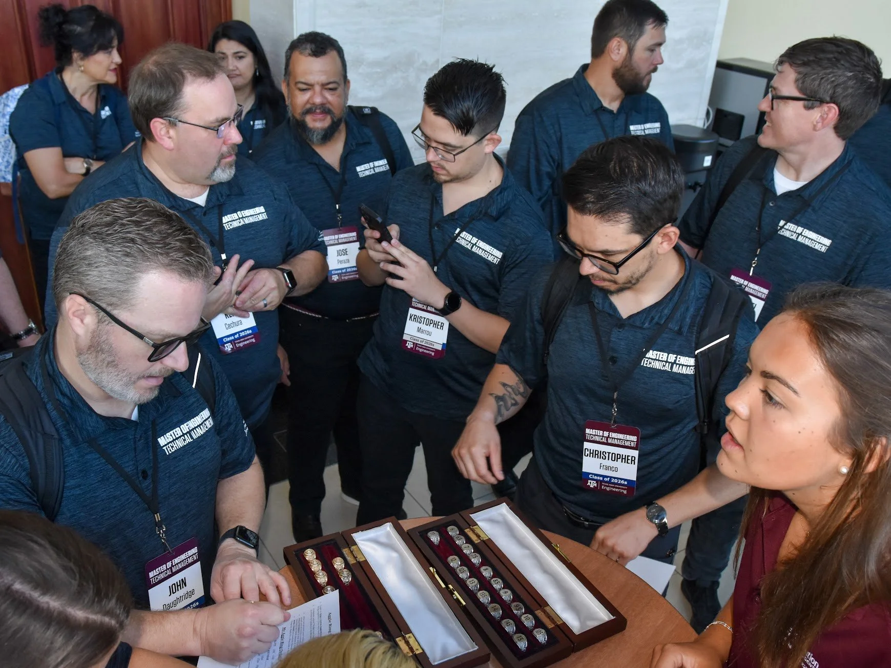Group of people gathered around a table looking at and discussing a box of medals or awards, with some individuals holding documents and one person using a smartphone, all wearing matching navy blue shirts with name tags.