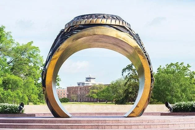 Large sculpture of a gold and black ring, forming an archway, situated in an outdoor park with trees and a building in the background.