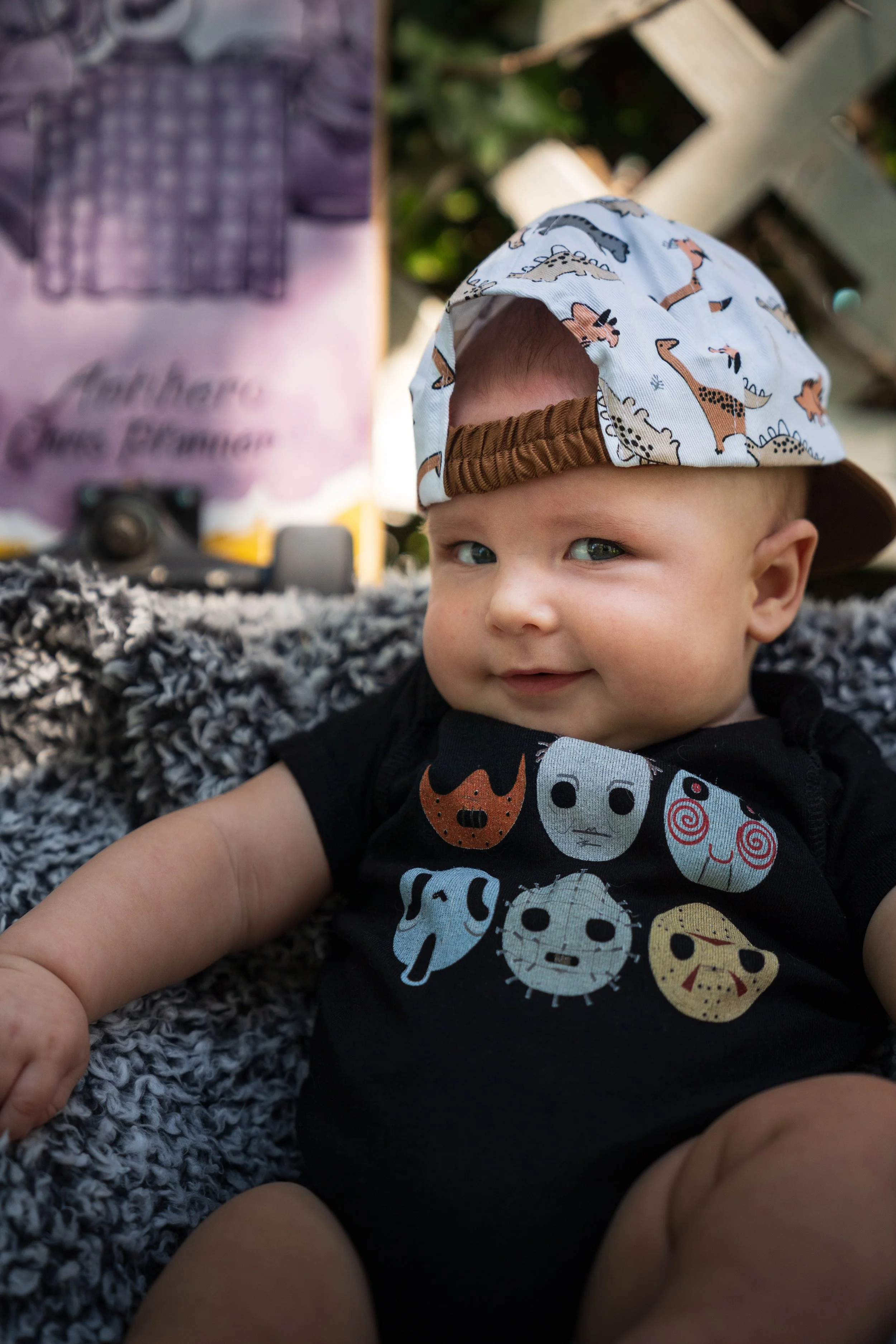 A baby boy with blue eyes wearing a cap with dinosaur prints and a black shirt with monster faces, sitting on a textured blanket outdoors.