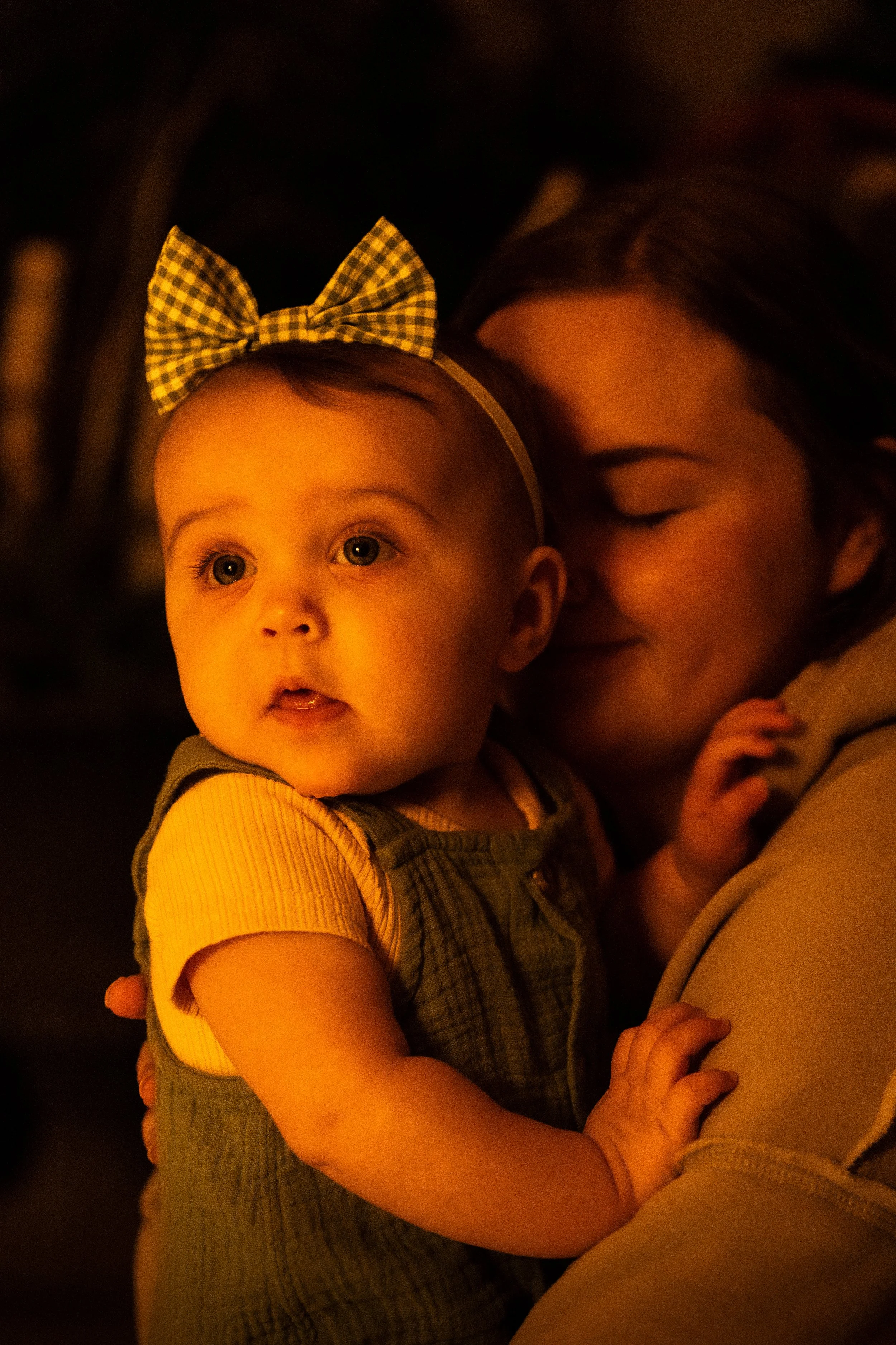 A young girl with a checkered bow on her head is being held by a her mother, both with warm lighting highlighting their faces, in a close-up shot showing affection and warmth.