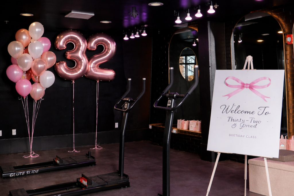 Pink and white balloons, birthday number balloons '32', a welcome sign with pink bow and text for a thirty-two birthday celebration, exercise equipment, and gift bags in the background.