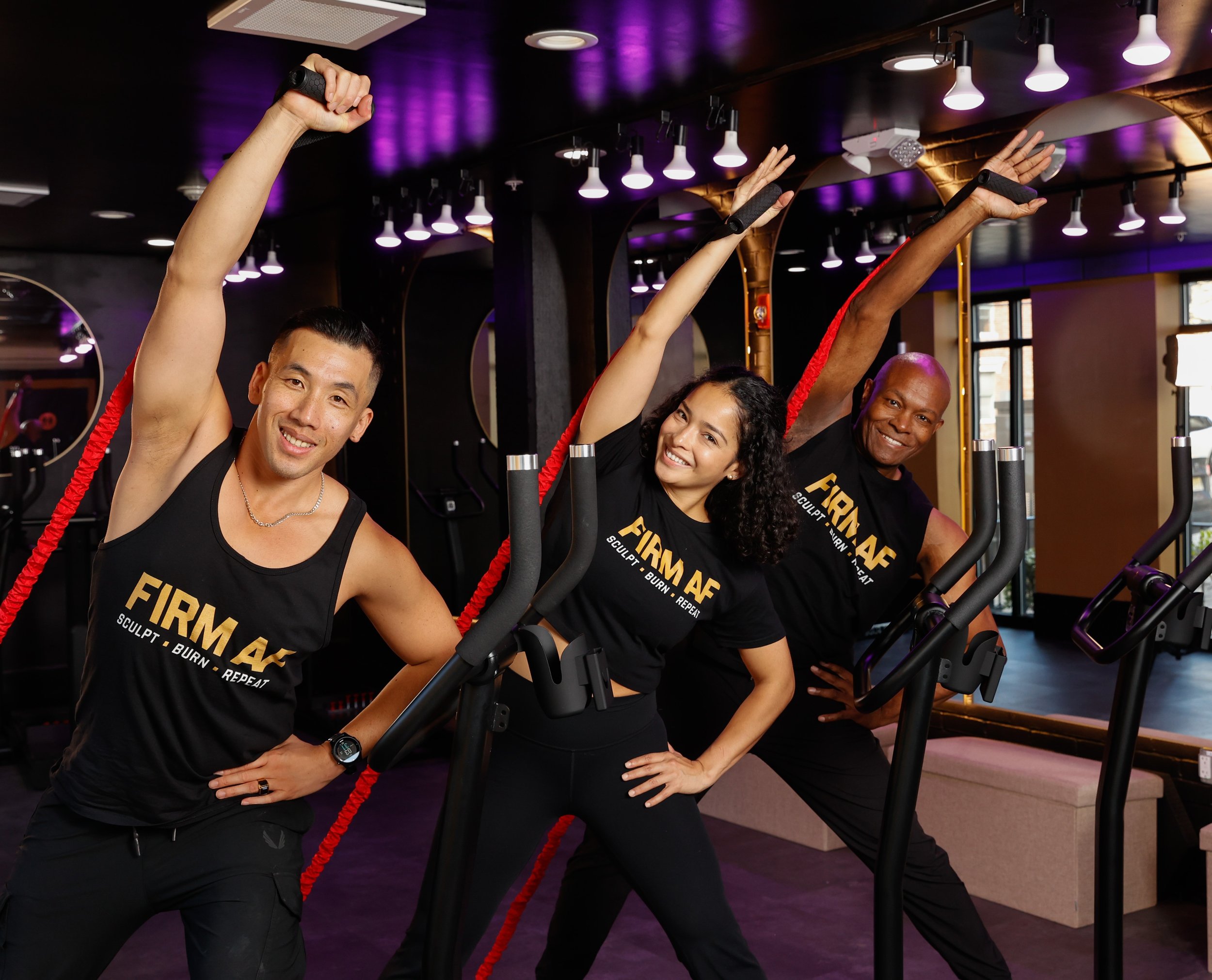 Three people in a gym posing happily with stretching poses, wearing matching black shirts with the phrase 'FIRM AF' on them.