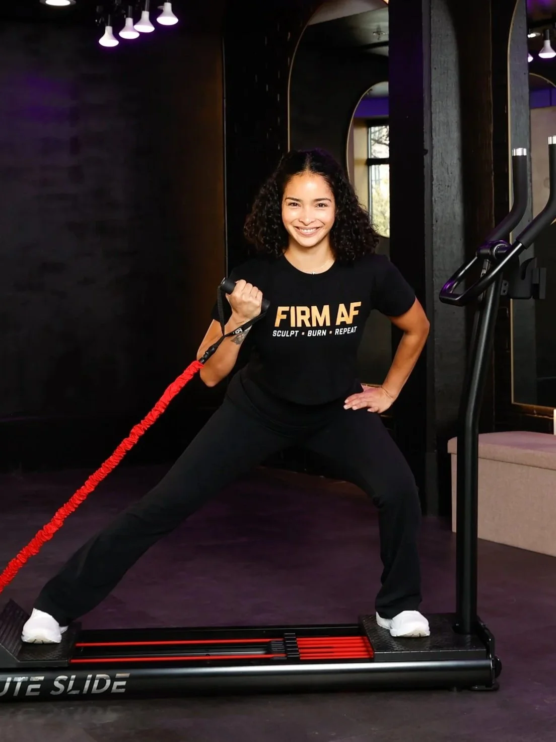 Jessica, a latin woman, in black athletic clothes smiling, holding a fitness resistance band, standing on a Glute Slide Machine in a FIRM AF fitness studio.