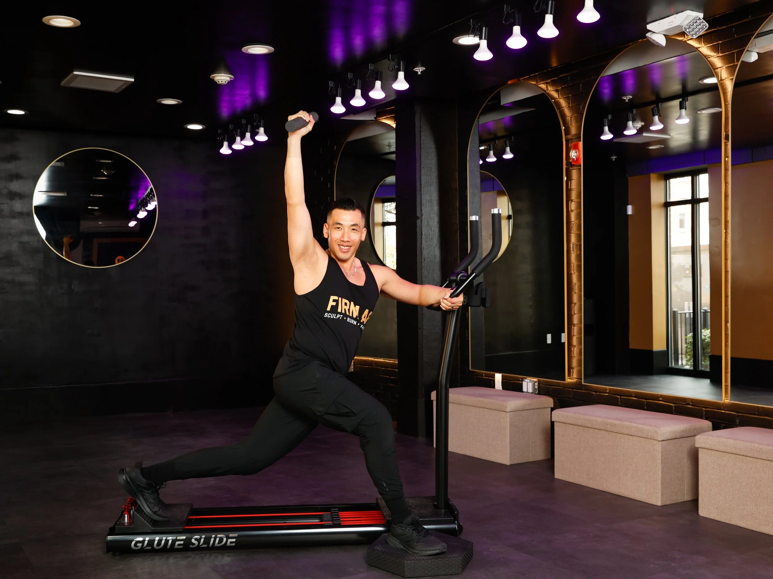 Sonny, an Asian man, exercising on a Glute Slide machine in FIRM AF fitness studio, holding a small weight in one hand and one hand on the Glute Slide Machine, smiling, with purple ceiling lights and large mirrors in the background.