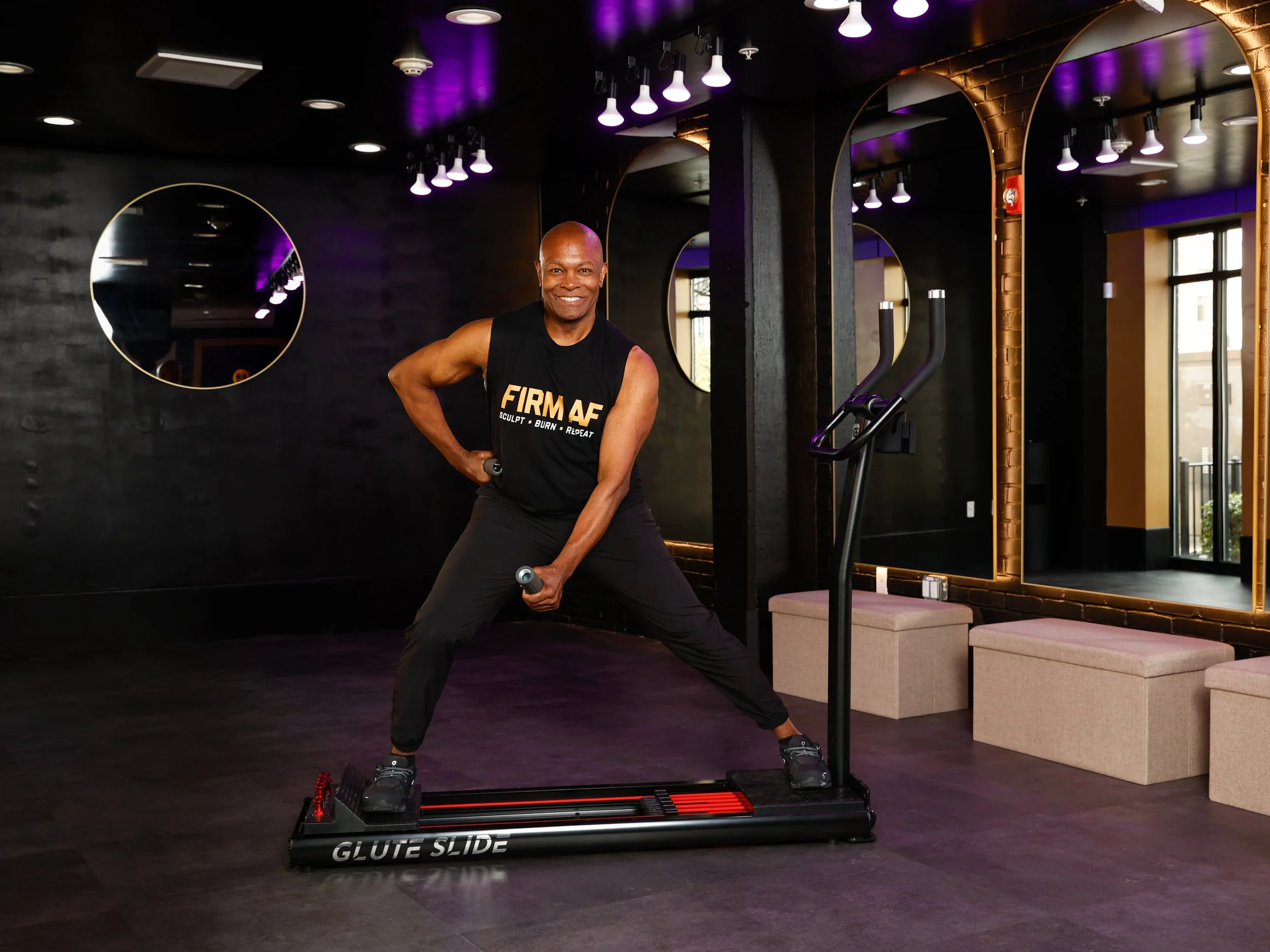 Carl, a black man, smiling in black workout clothes holding small dumbbells on a Glide Slide Machine in a FIRM AF fitness studio with mirrors, purple lighting, and seating area