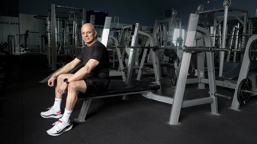 Man sitting on gym bench in a weightlifting gym with barbells and equipment in the background.