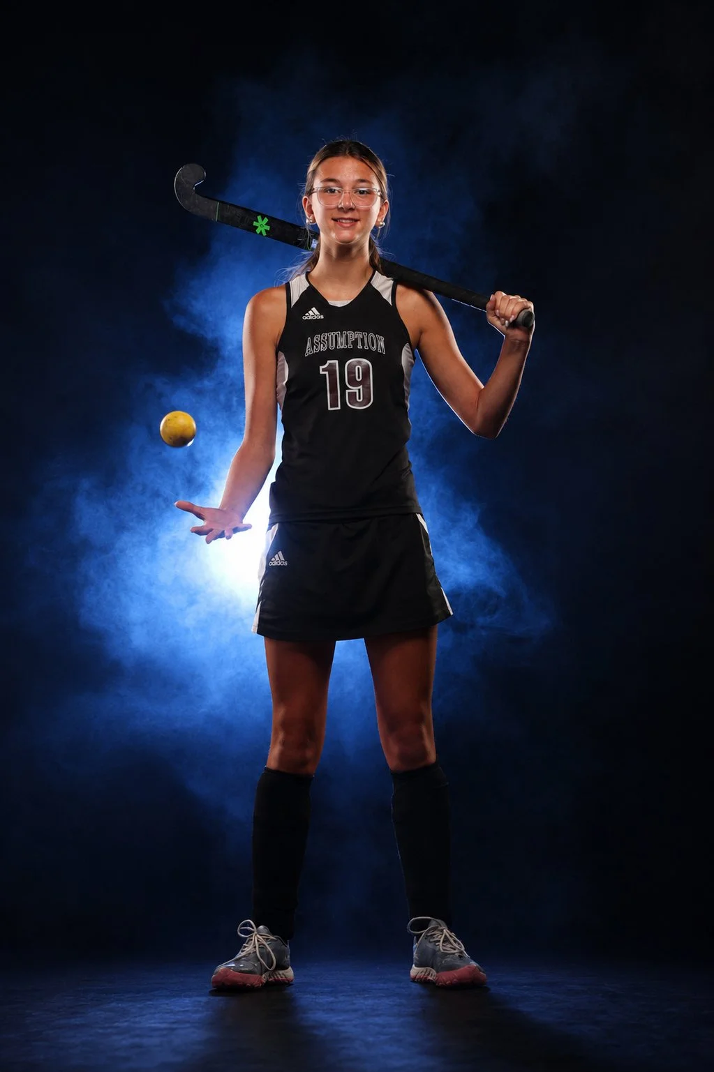 A young female field hockey player in a black uniform with the number 19, holding a field hockey stick on her shoulder, standing in a dark studio with a blue backlight and fog, with a yellow ball in mid-air nearby.