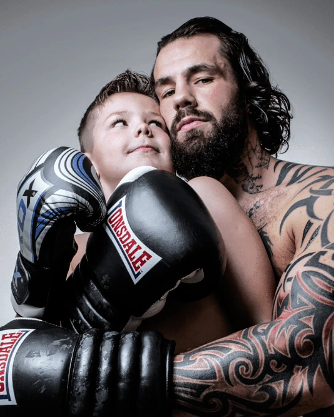 A tattooed man with long dark hair and a beard holds a young boy with short hair, both wearing boxing gloves, close together in a pose with serious expressions.
