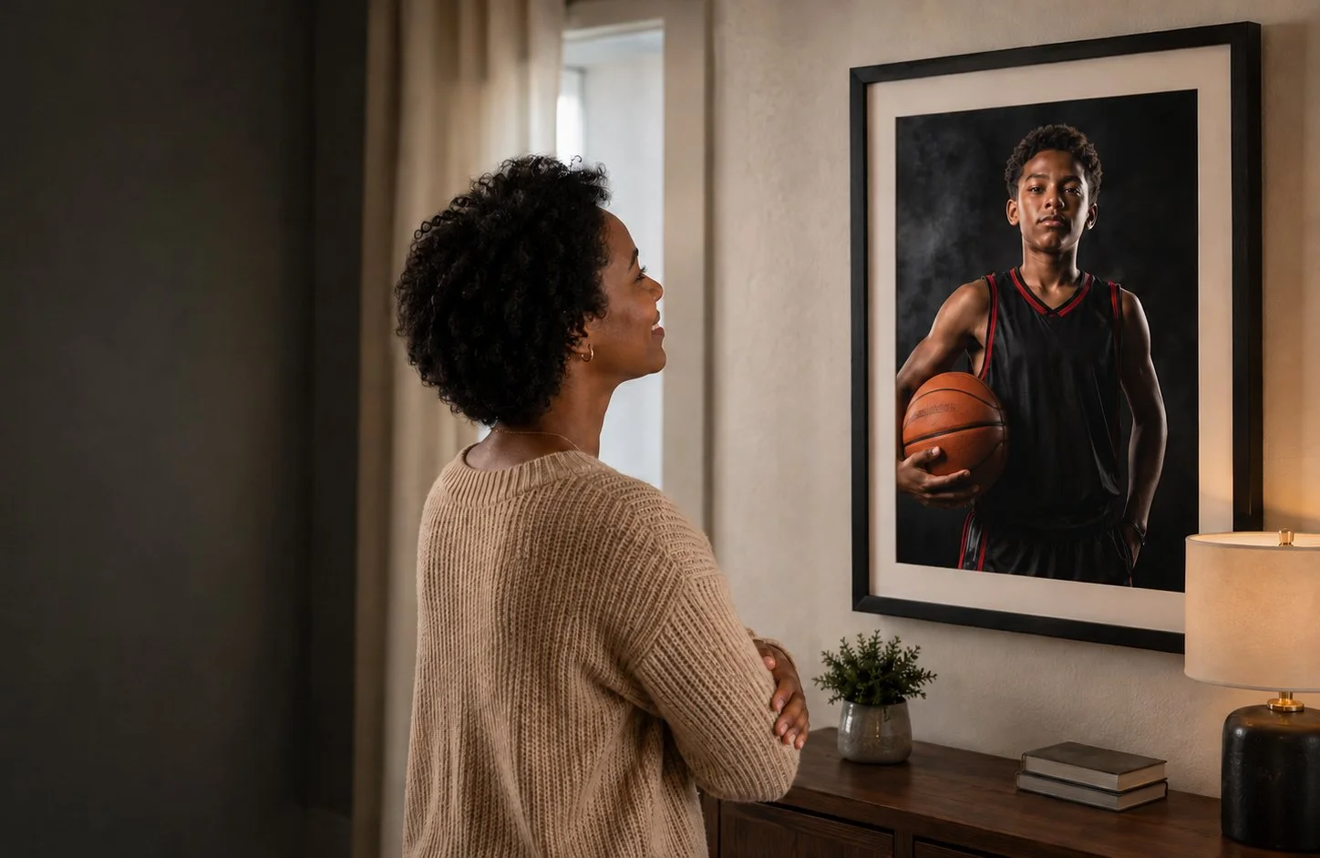 A woman with curly hair and a beige sweater smiling while looking at a large framed photograph of a young basketball player holding a basketball.
