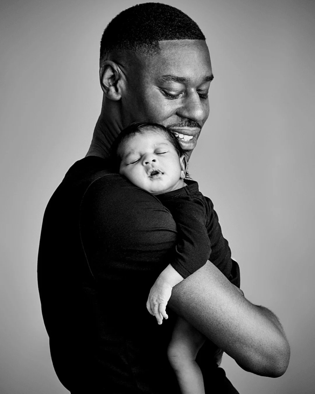 A black and white photo of a man holding a sleeping baby close to his shoulder, both smiling gently.