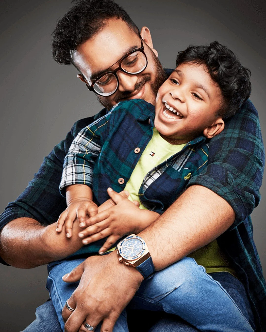 A joyful young boy hugging a smiling man with a beard and glasses, both wearing casual plaid shirts and blue jeans, against a neutral gray background.