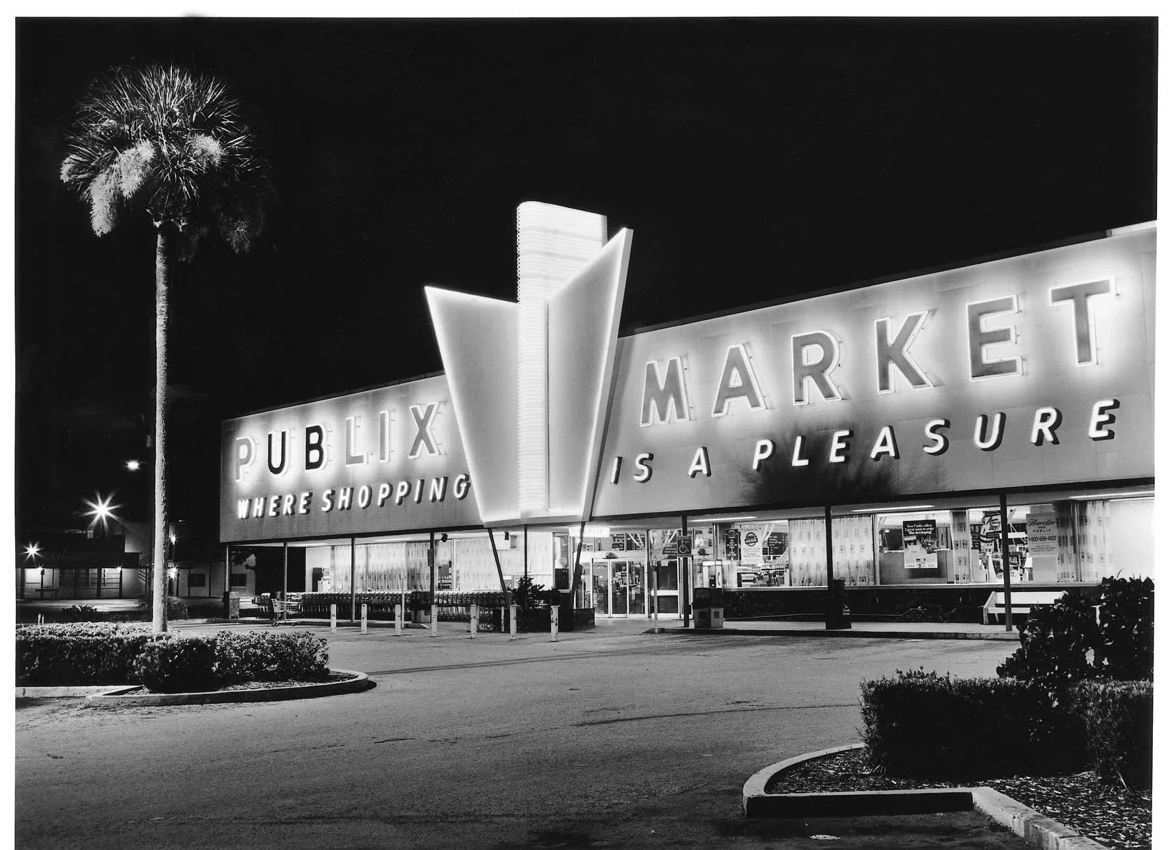 Black and white photo of a Publix grocery store at night with illuminated sign reading "Publix Market is a Pleasure" and a tall palm tree on the left side.