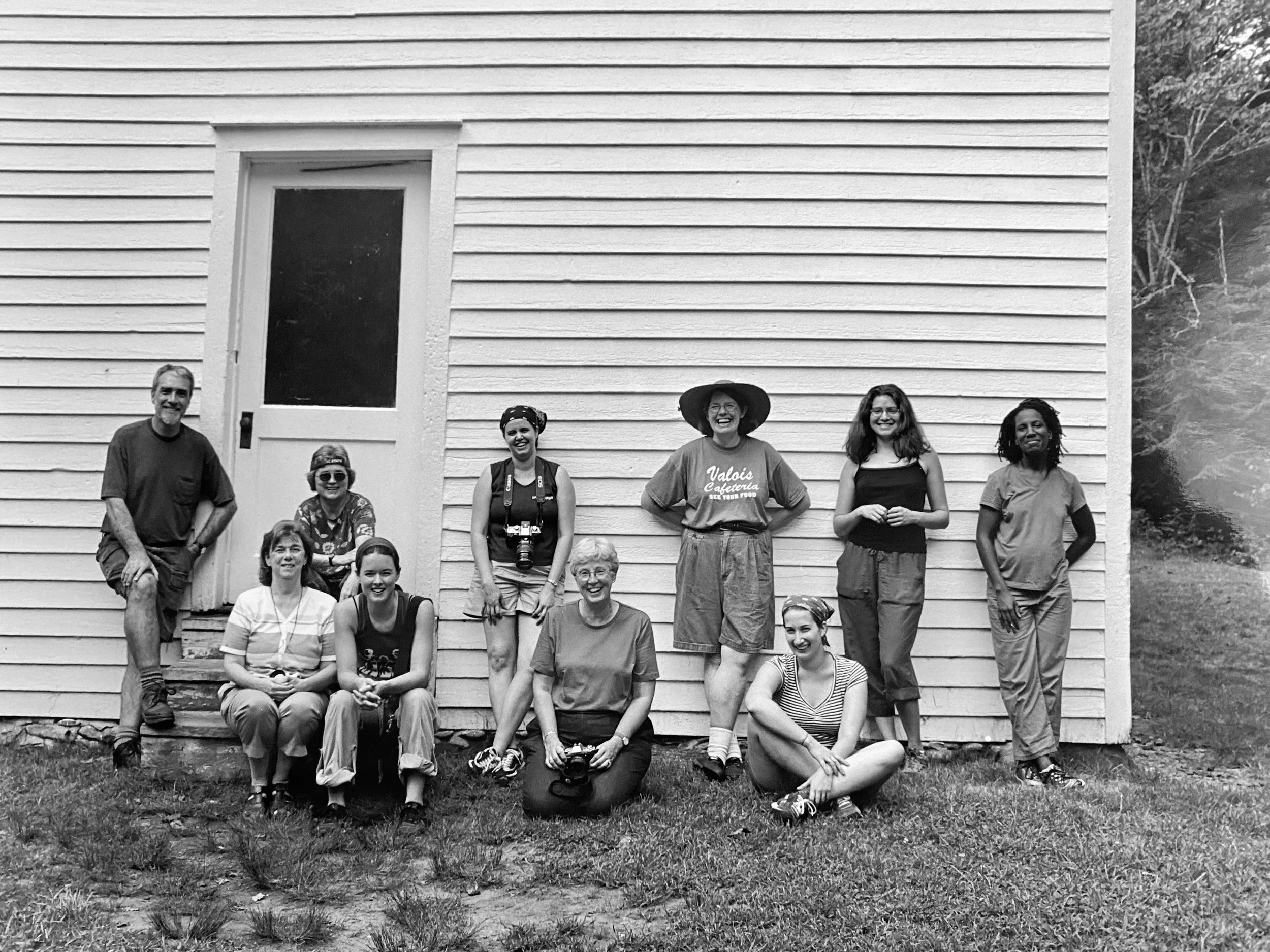Black and white photo of a group of approximately 13 people standing and sitting in front of a white wooden building, smiling for the camera.