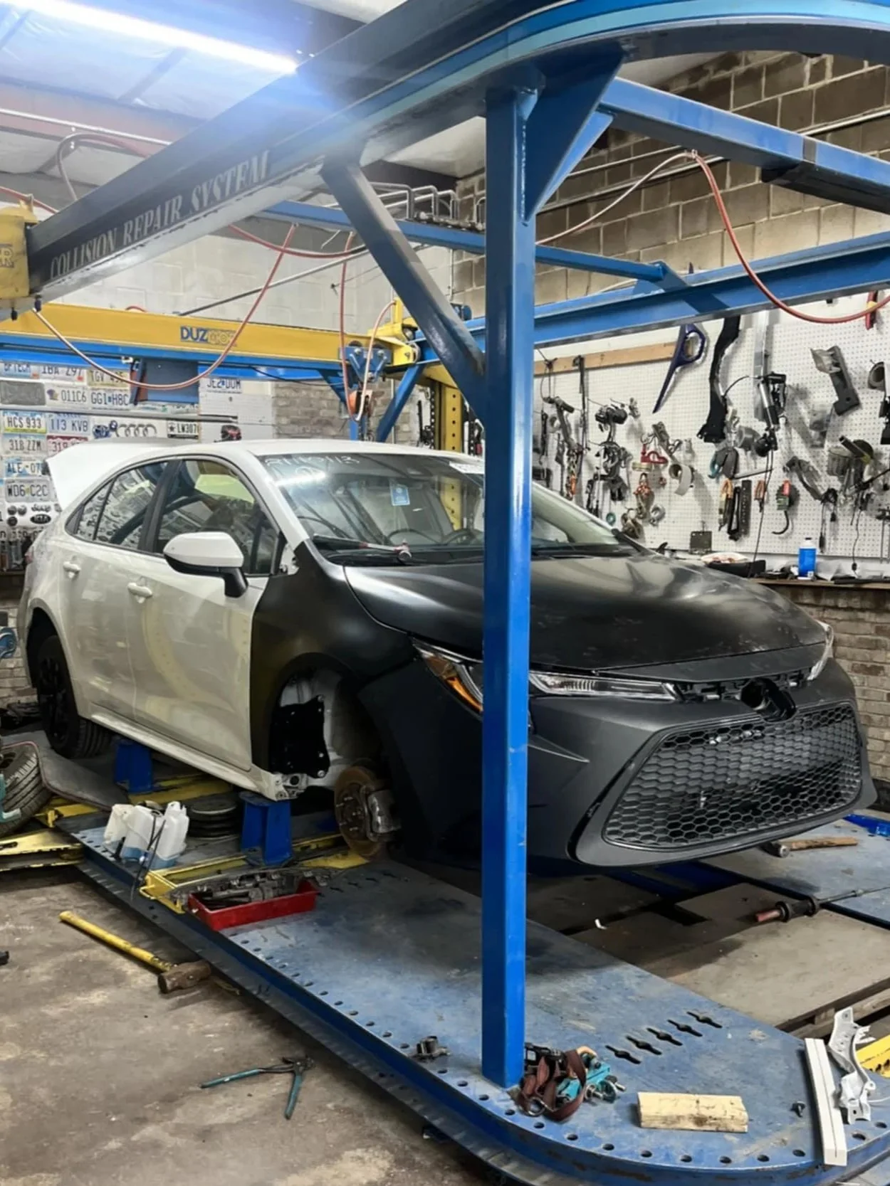 A white and black sedan, possibly a Honda Civic, is in a repair shop on a lift with front wheels removed. The repair shop has tools hanging on the wall and license plates displayed in the background.