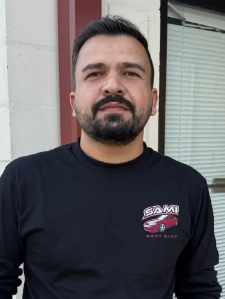A man standing outside in front of a brick and concrete wall, wearing a black long sleeve shirt with a logo of a red car and the words 'SAMI Body Shop'.