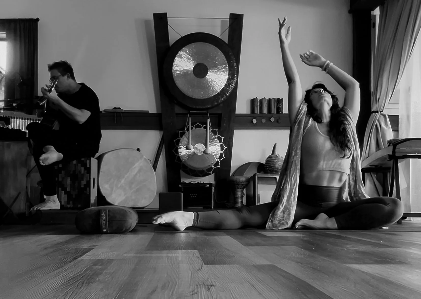 Kaitana Magno & Jimmy Sutherland lead Sonic Soma. Kaitana is practicing yoga in a split position with arms raised inside a room, while a man plays guitar in the background.