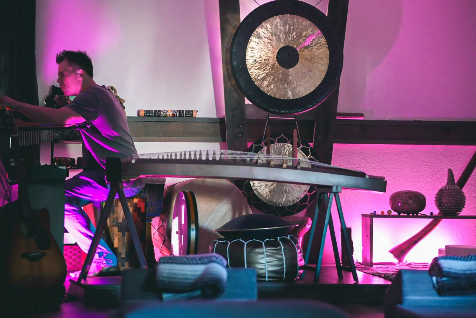 A musician playing keyboard on stage with pink and purple lighting, surrounded by various musical instruments, a gong, a guitar, and decorative objects.
