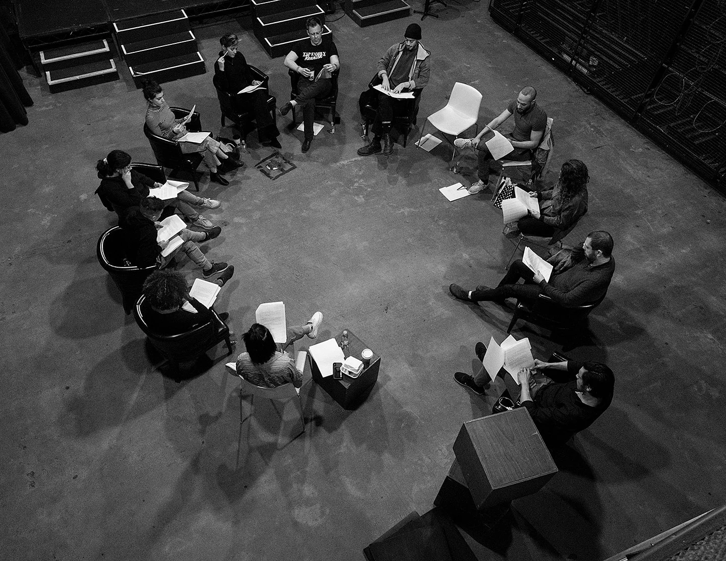 Immersive workshop. Kaitana Magno & Jimmy Sutherland lead a group of people seated in a circle, reading scripts or papers, in a studio or rehearsal space with some stairs and equipment visible.