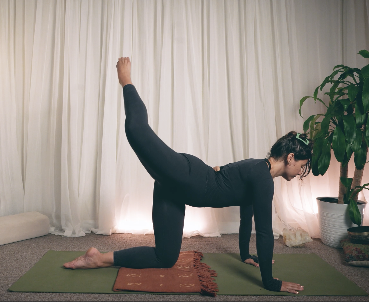 Kaitana Magno practicing yoga in a room with white curtains, a potted plant, and warm lighting. She is on a green yoga mat, wearing black leggings and a black long-sleeve top, performing a yoga pose.