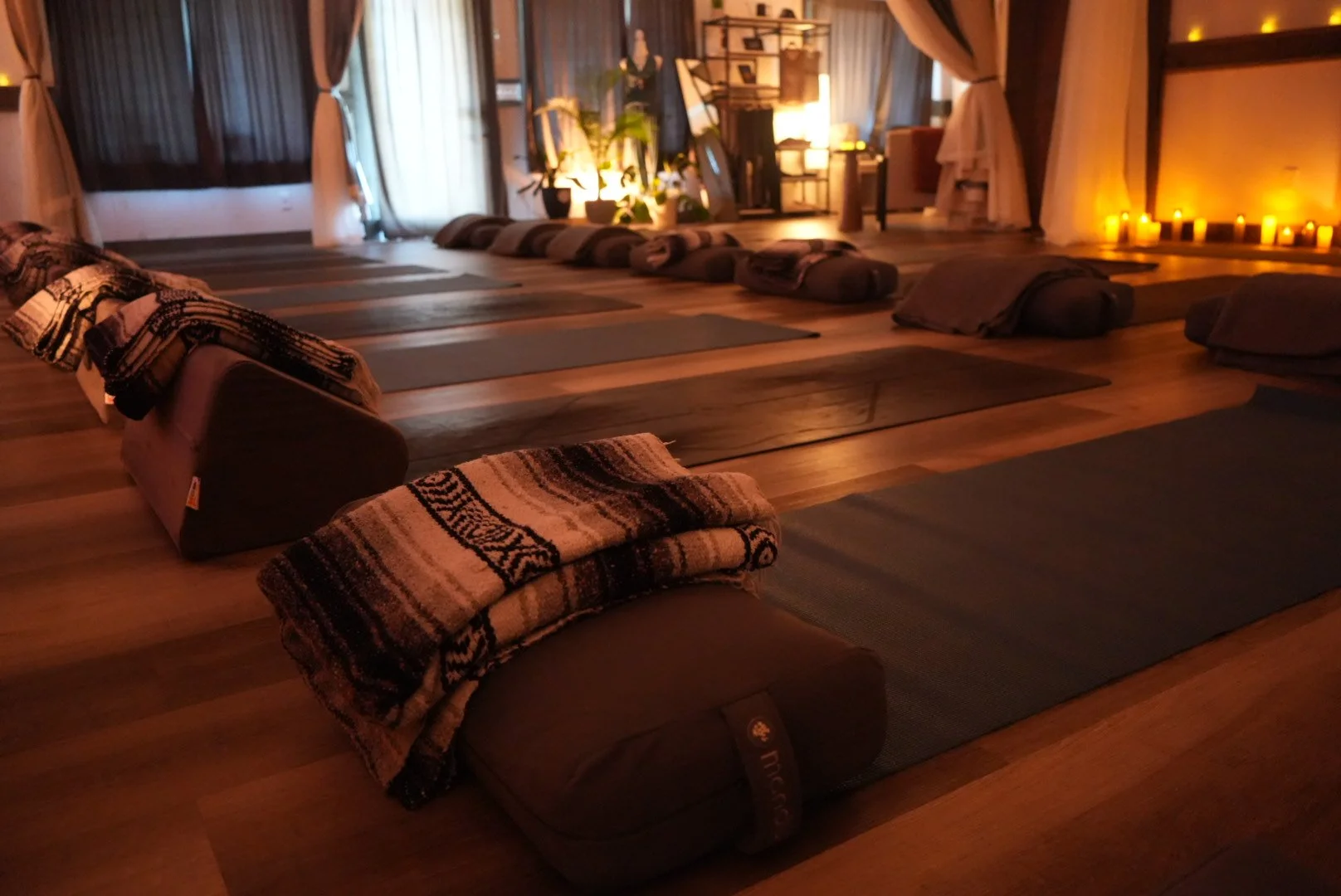 Yoga mats and bolsters arranged on a wooden floor in a dimly lit room, with candles along the walls and curtains on large windows.