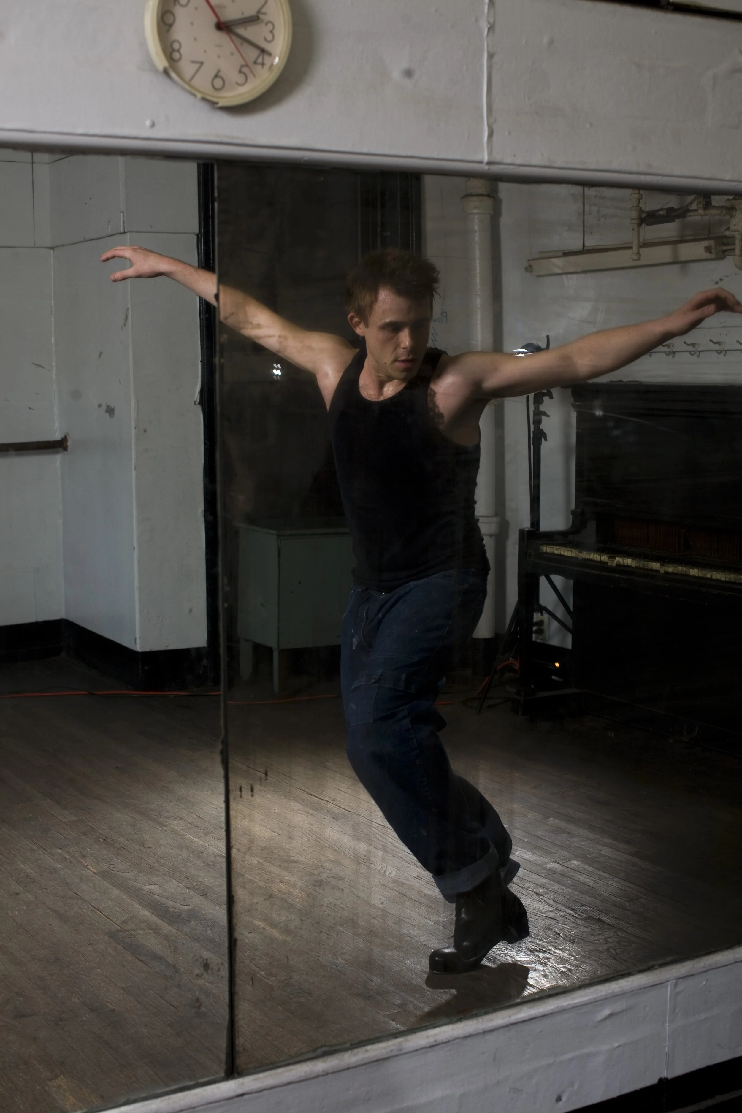 Jimmy TapZ Sutherland tap dancing in a black tank top and jeans dancing in Fazil's Time Square Studio on a wooden floor, reflected in a mirror, with a clock on the wall above him.