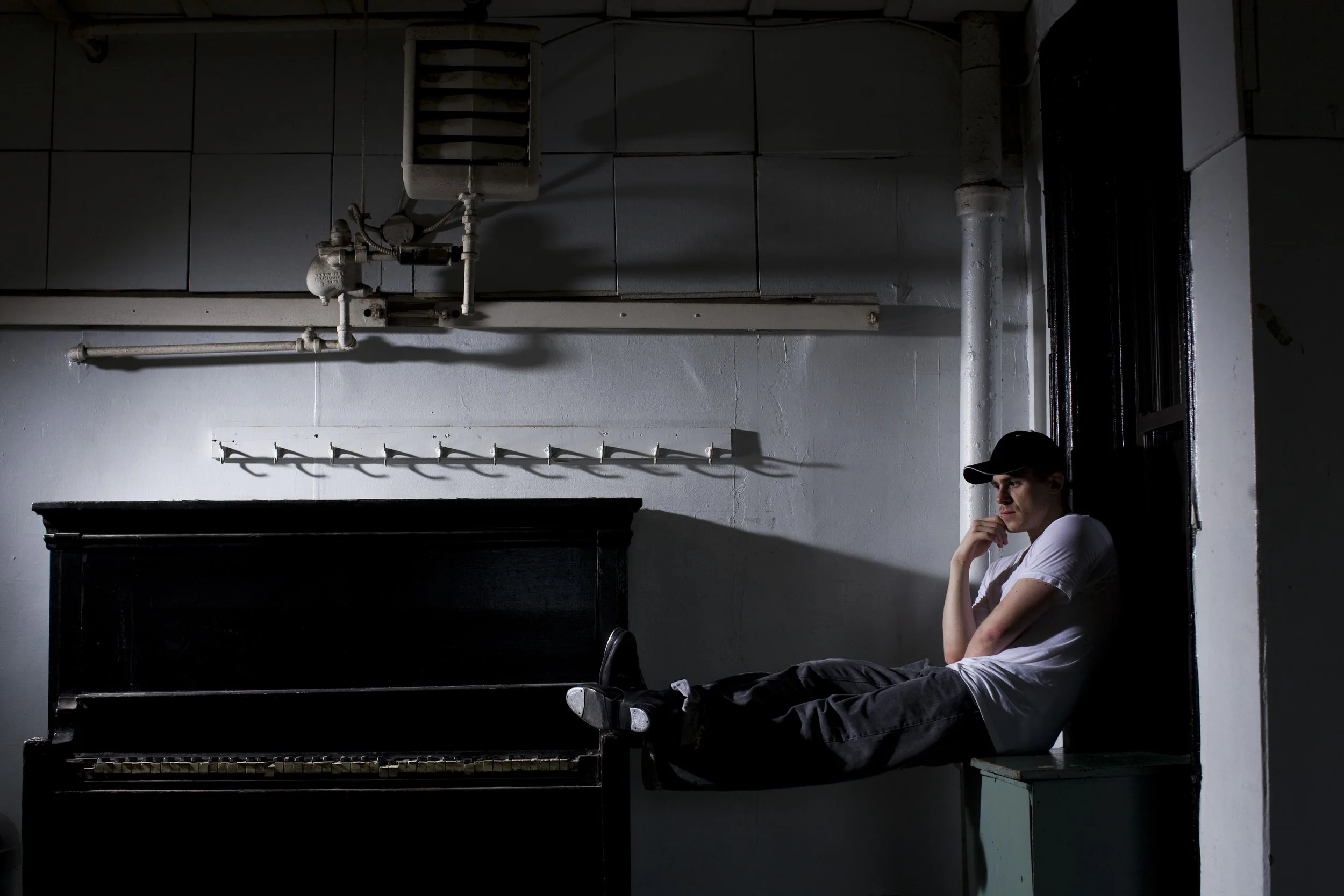 Jimmy TapZ Sutherland in Fazil's in NYC. Wearing a white t-shirt and black pants, wearing a baseball cap, sitting on a small ledge, resting his head on his hand, in a dimly lit room with an old black upright piano and industrial pipes on the wall.