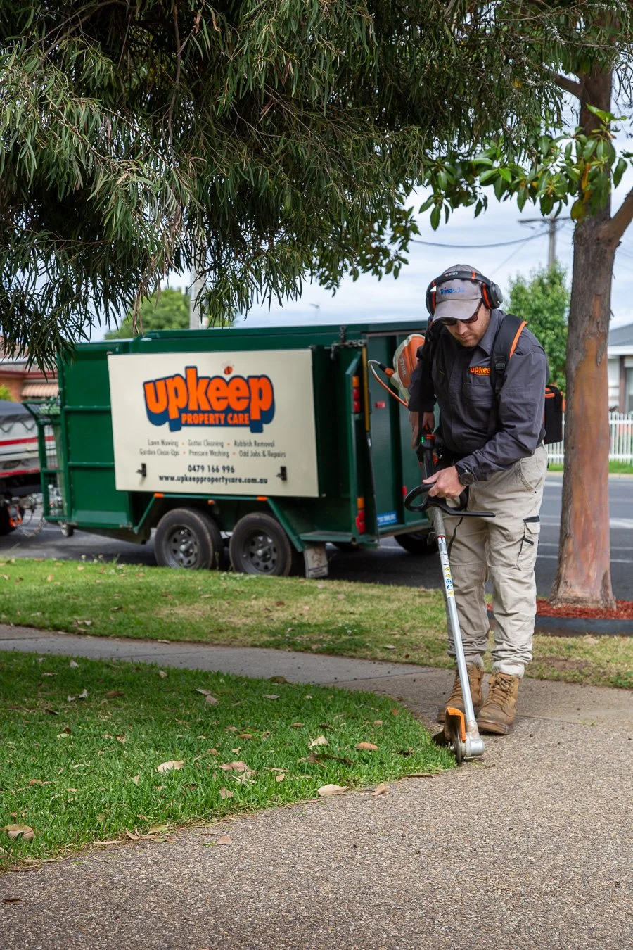 A worker using a grass trimmer on a footpath near a tree, with a green property care truck in the background.