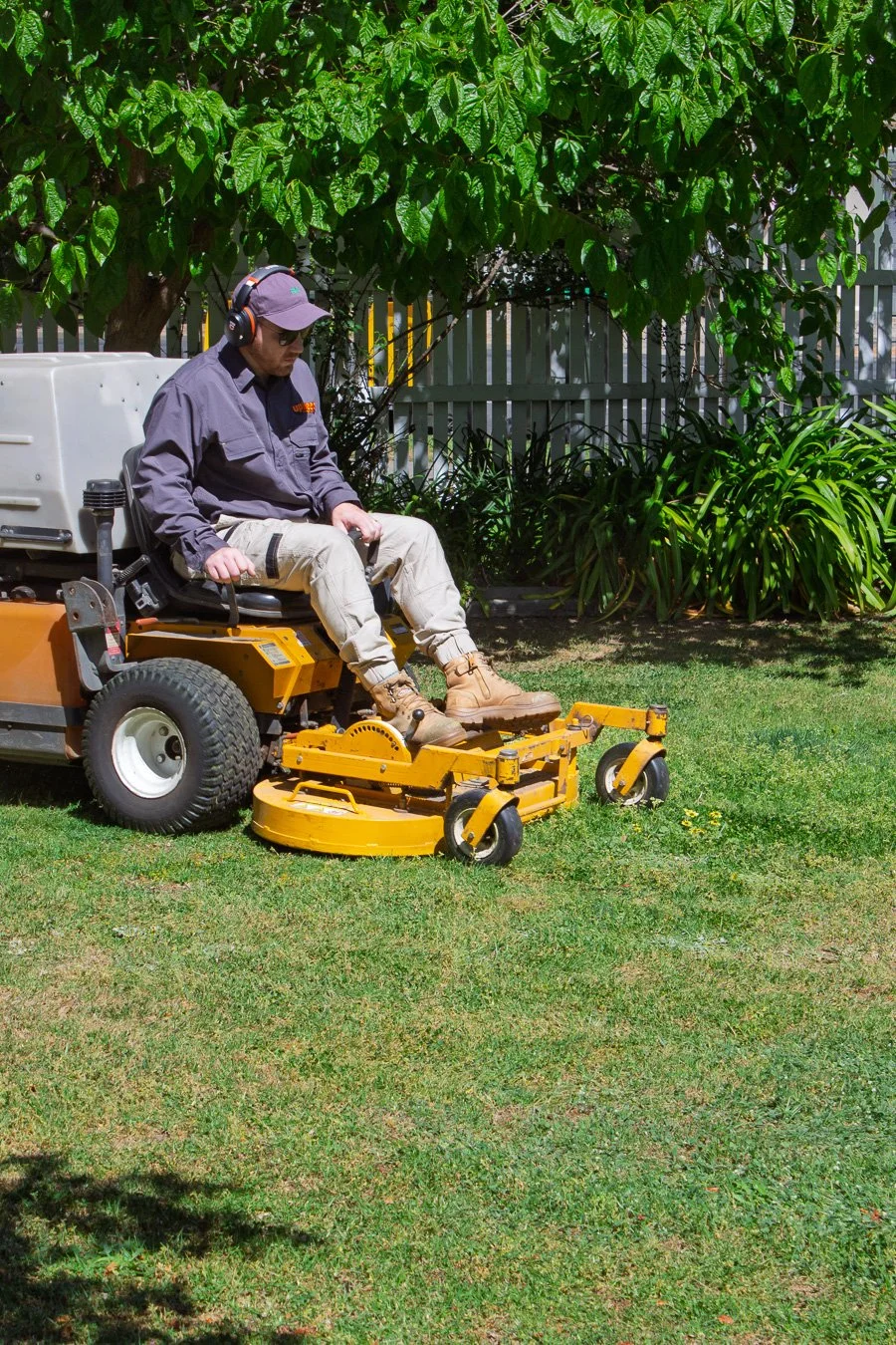 A person riding a yellow riding lawn mower on a lawn, with a tree, bush, and fence in the background.