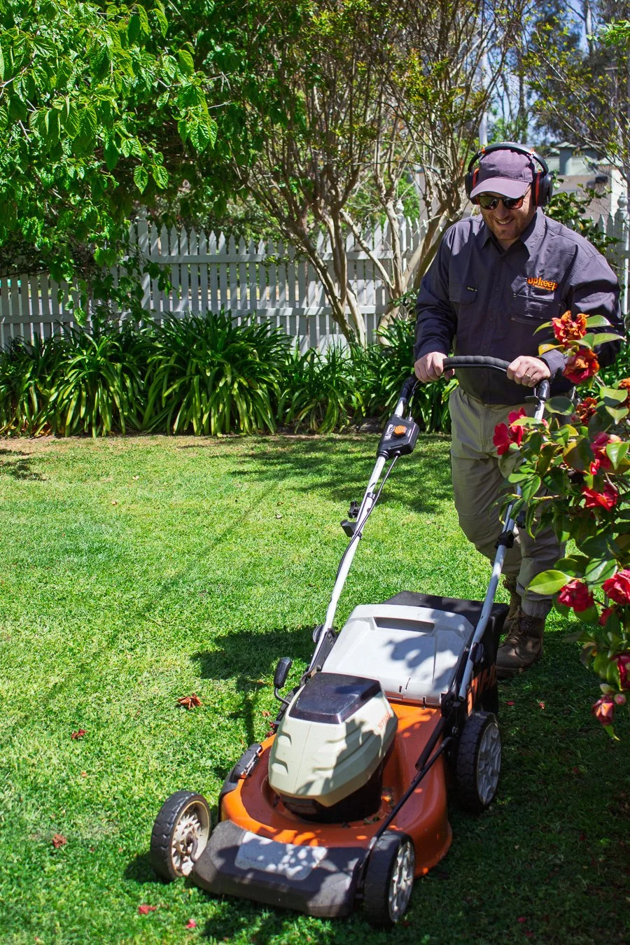 A man using a lawn mower in a backyard with green grass, bushes, trees, and a white fence in the background on a sunny day.