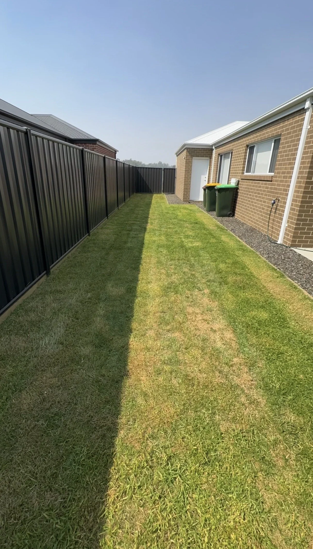 A tidy backyard with a narrow strip of green grass, bordered by a black metal fence on the left and a brick house with two green garbage bins on the right, under a clear blue sky.