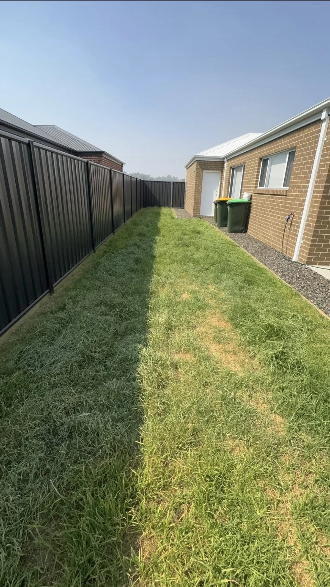 Backyard with grass lawn, black metal fence on the left, brick house on the right, window, door, and two green trash bins.