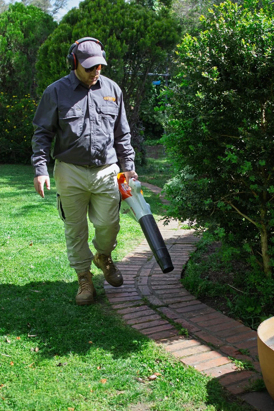 Man in outdoor clothing using leaf blower to clear a brick path, surrounded by green grass and bushes.
