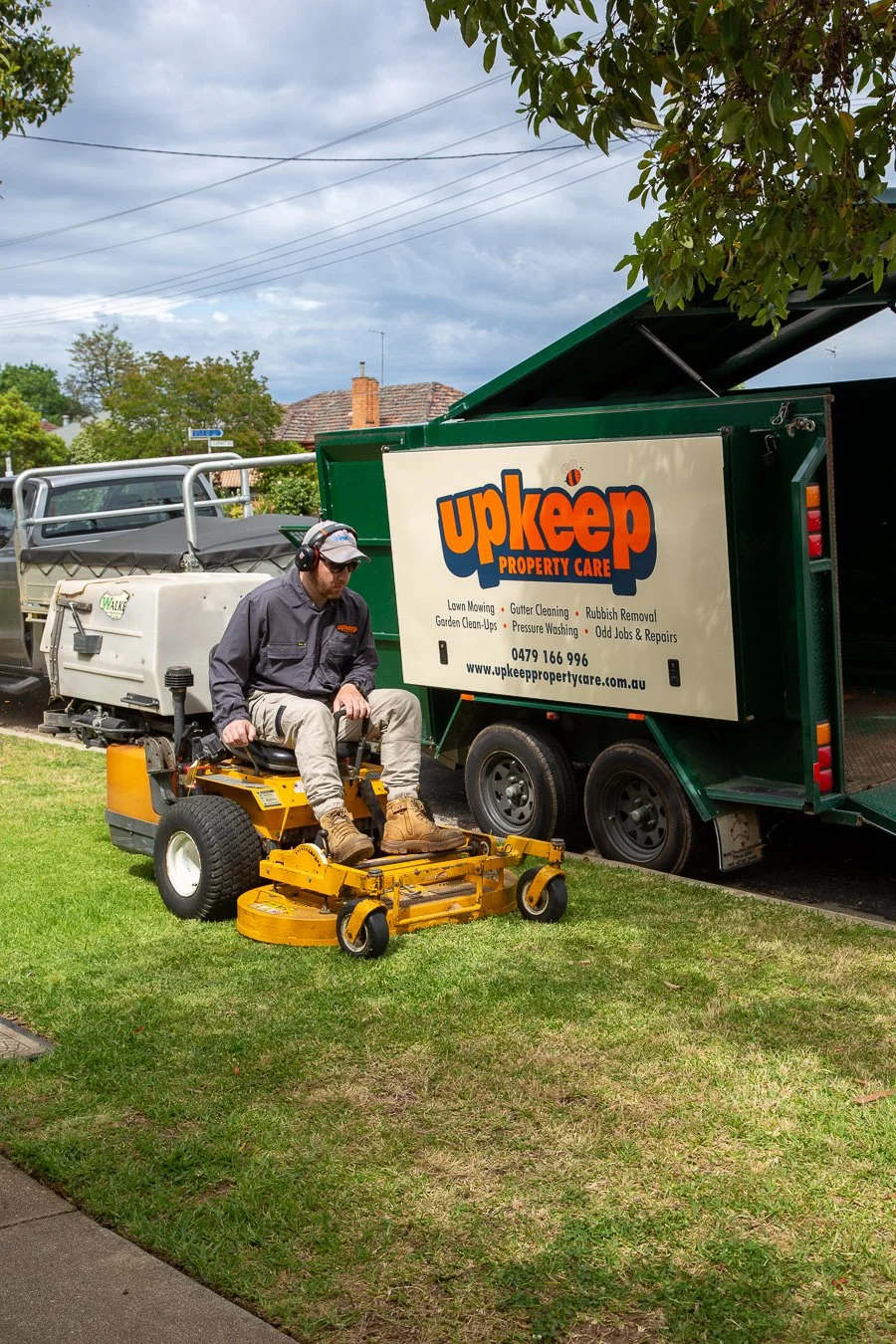A man in a gray uniform and cap operating a yellow ride-on lawn mower on a grassy lawn, near a green Upeep Property Care truck with an open side door, under a cloudy sky in a residential neighborhood.