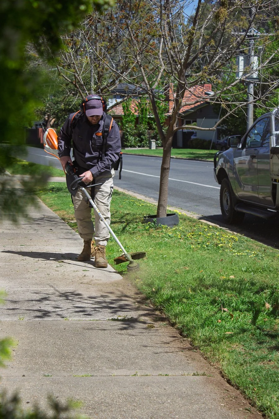 A man wearing a cap, headphones, and outdoor work gear is trimming the sidewalk with a weed trimmer on a sunny day in a suburban neighborhood.