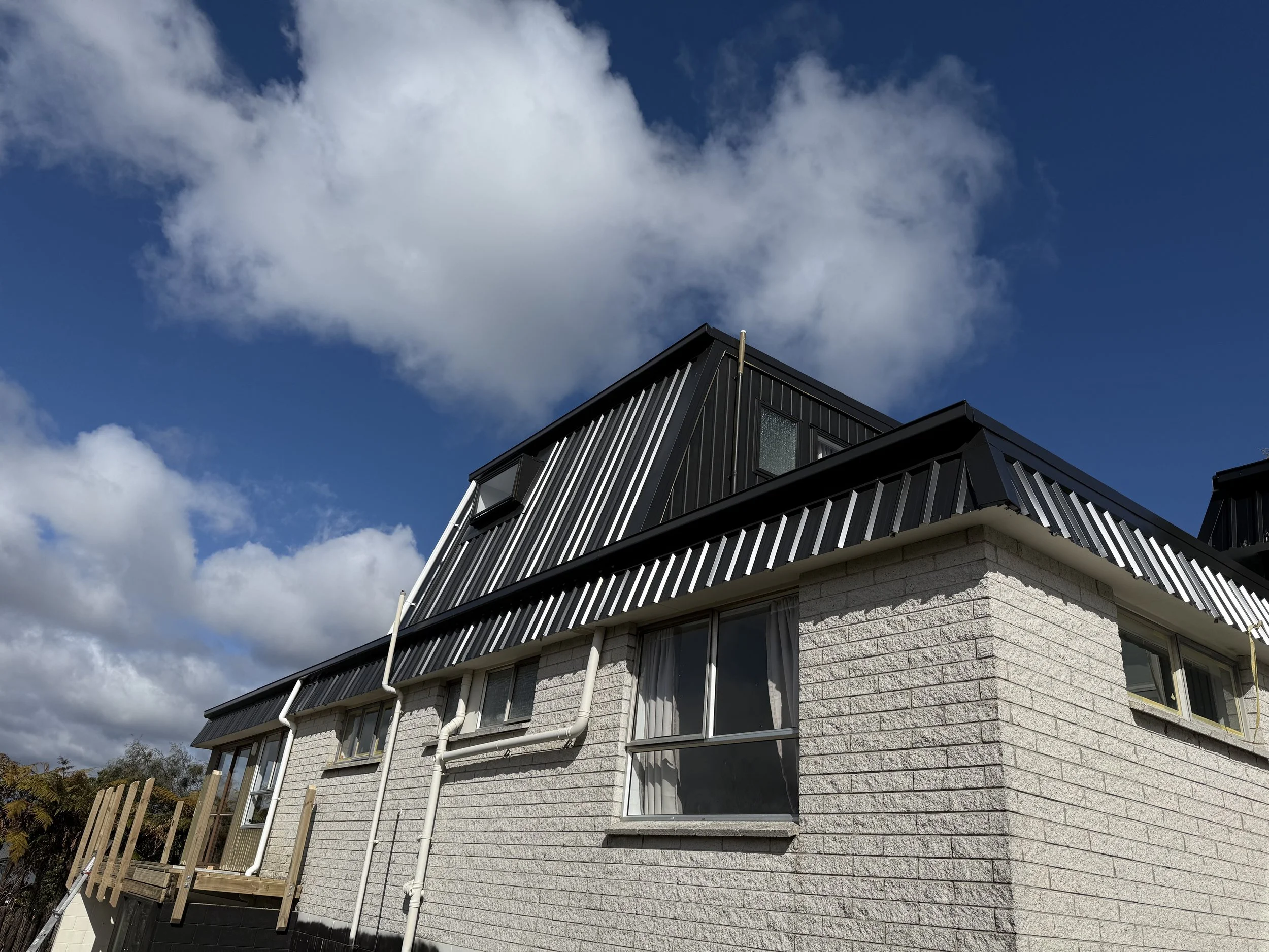A multi-story house with white brick walls, black metal roofing, and large windows, under a blue sky with white clouds.