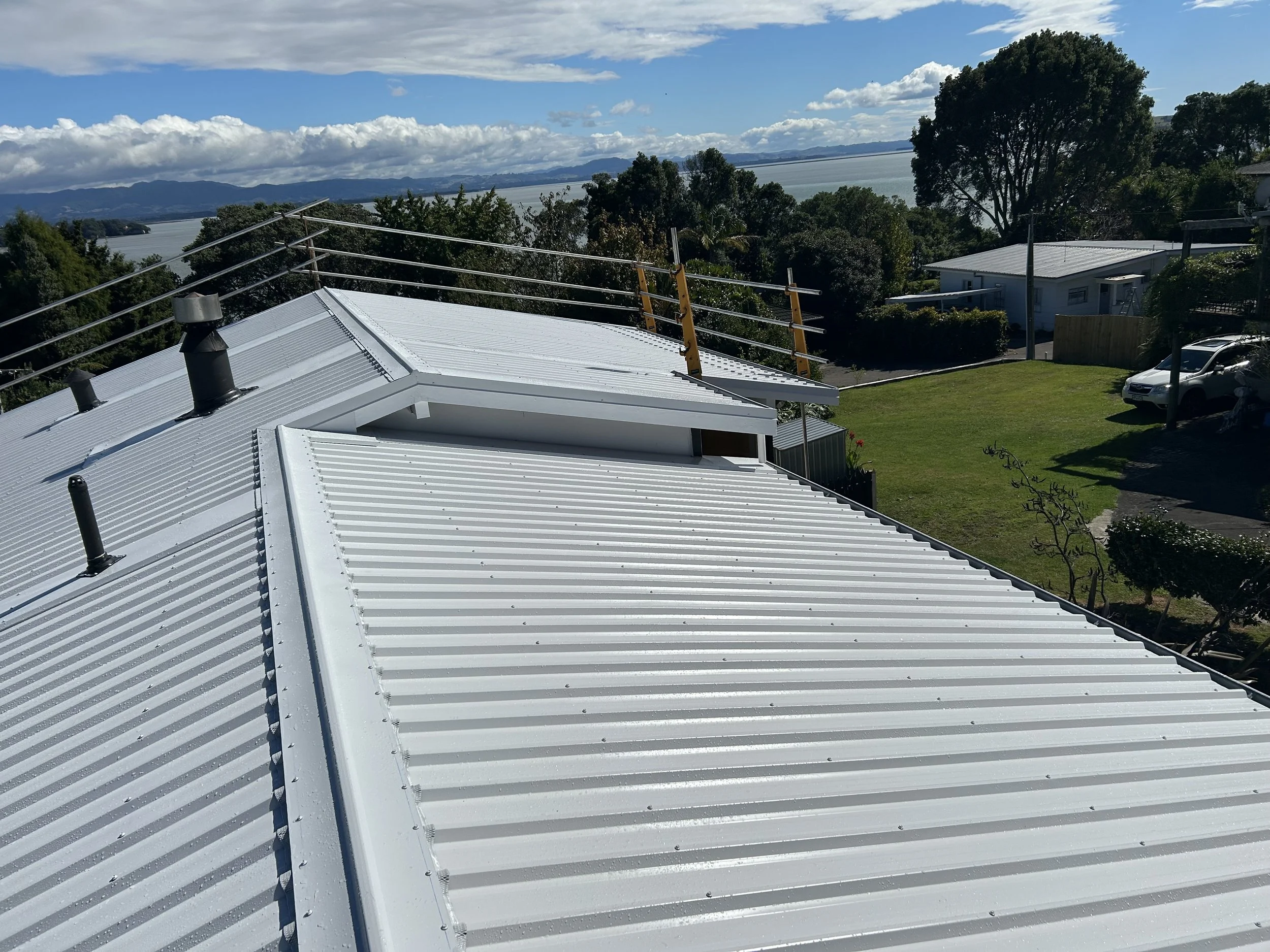 View of a white metal roof with vents and safety railings, overlooking a grassy yard, trees, a driveway with parked cars, and a body of water with mountains in the distance.