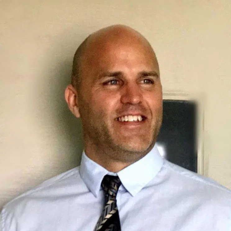 A person, smiling and wearing a white dress shirt with a patterned tie, standing against a light-colored wall.