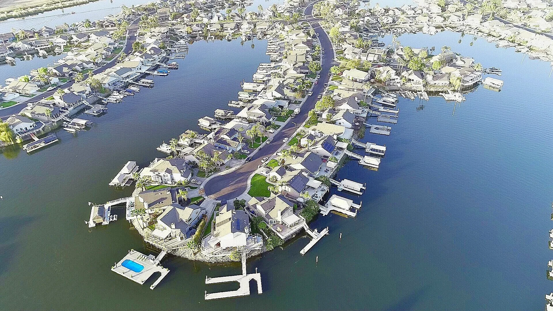 Aerial view of a residential neighborhood with houses along a waterway, boats docked at private piers, and a winding road.